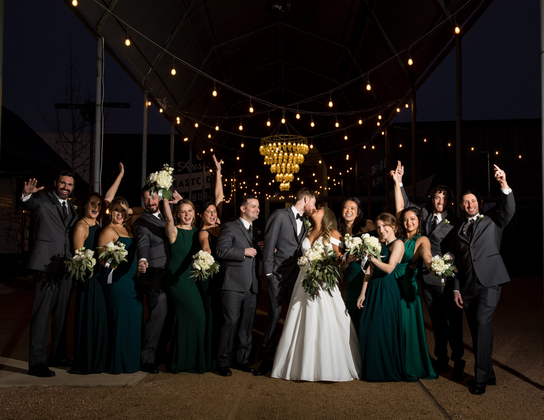 Bridal party celebrating under the Springdale Station chandelier — couple kissing in the center