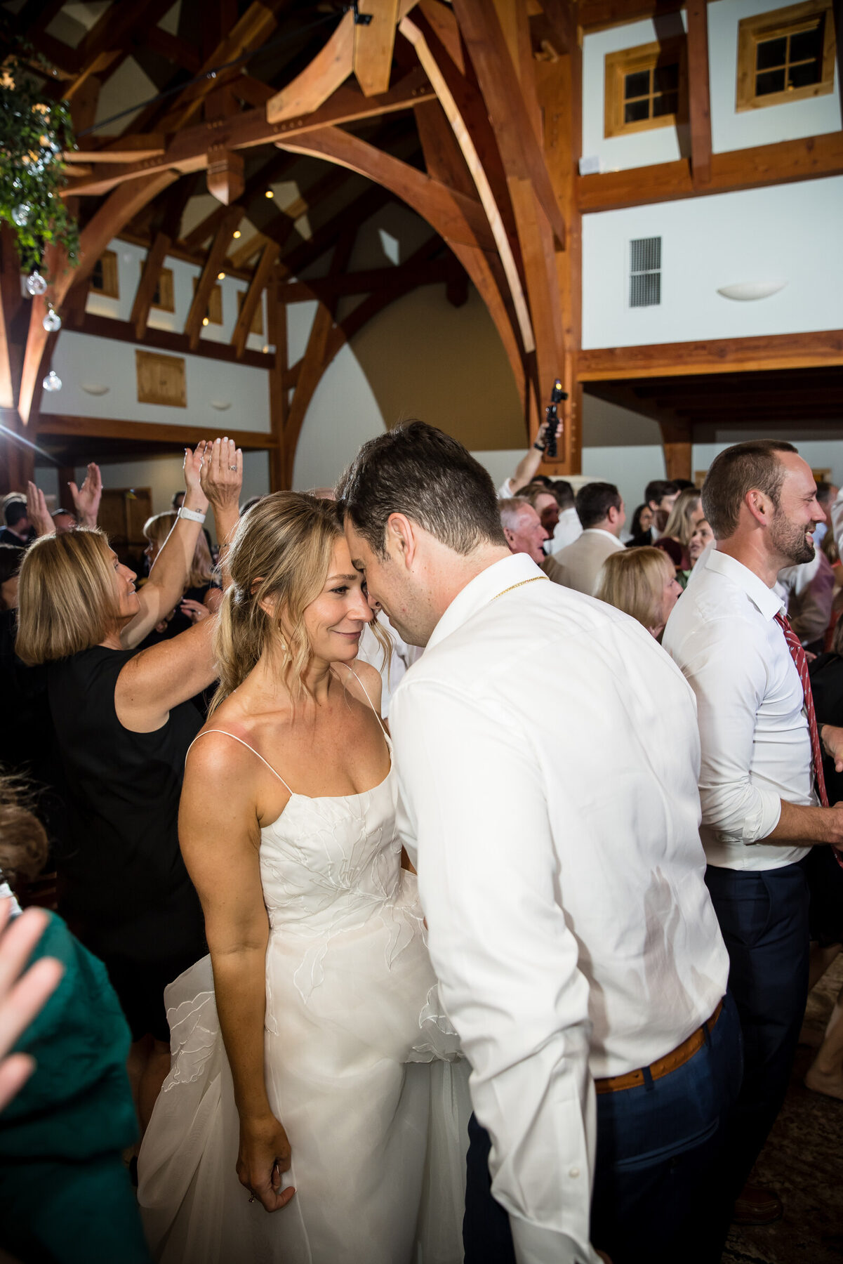 Dance floor candid — couple embracing in the middle of a packed reception under a chandelier, both laughing