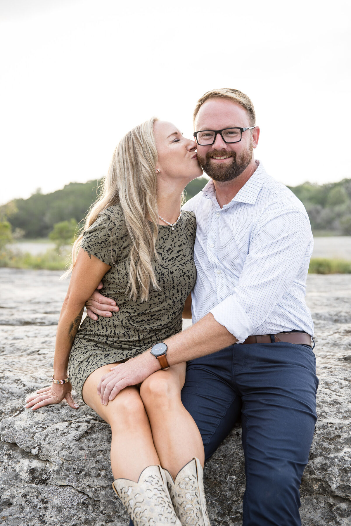 A couple sitting on a Hill Country rock outcrop at golden hour — woman in a green dress kissing her partner's cheek, cowboy boots, dusty pink Texas light