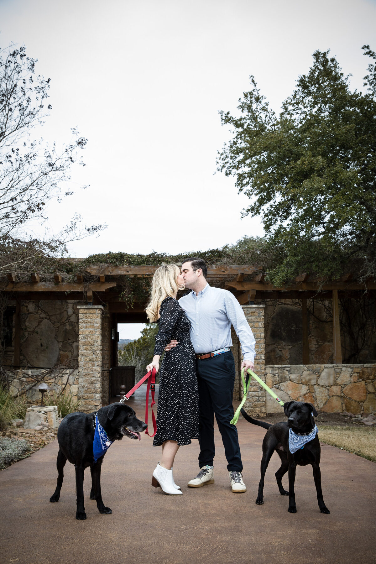 A couple kissing in front of a Hill Country stone archway with their two black Labs at their feet — a relaxed engagement session is the photographer's chance to fix camera-shyness before the wedding