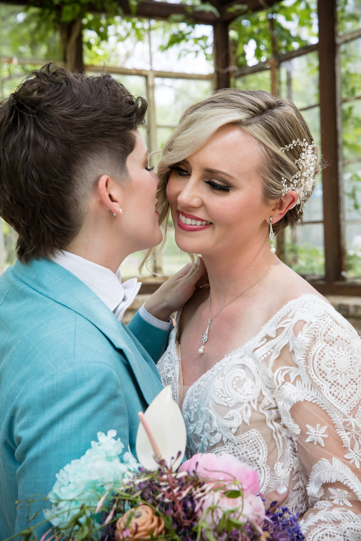 A queer couple sharing a tender greenhouse kiss — one in a teal three-piece suit, the other in an intricate lace gown — the kind of intimate candid The Bohde Collective is known for