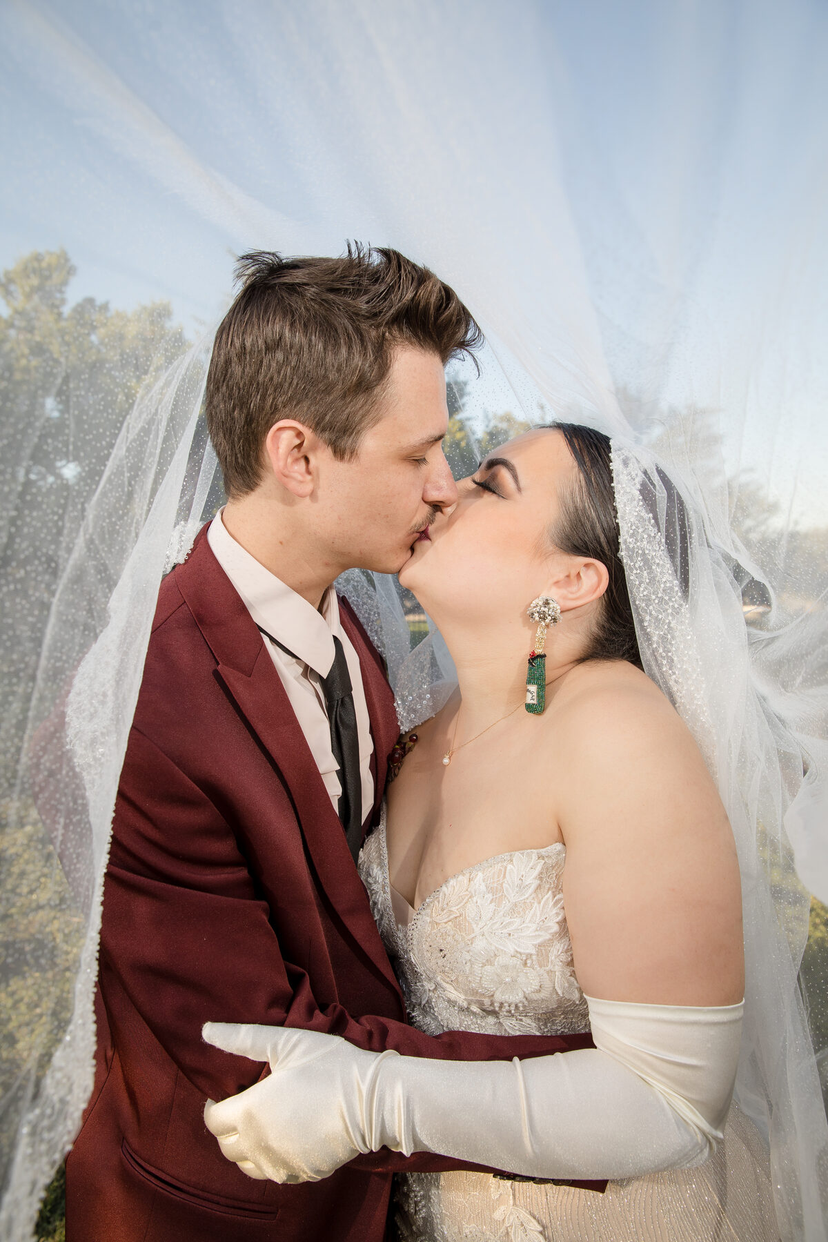 Couple kissing under veil, maroon suit and green earrings